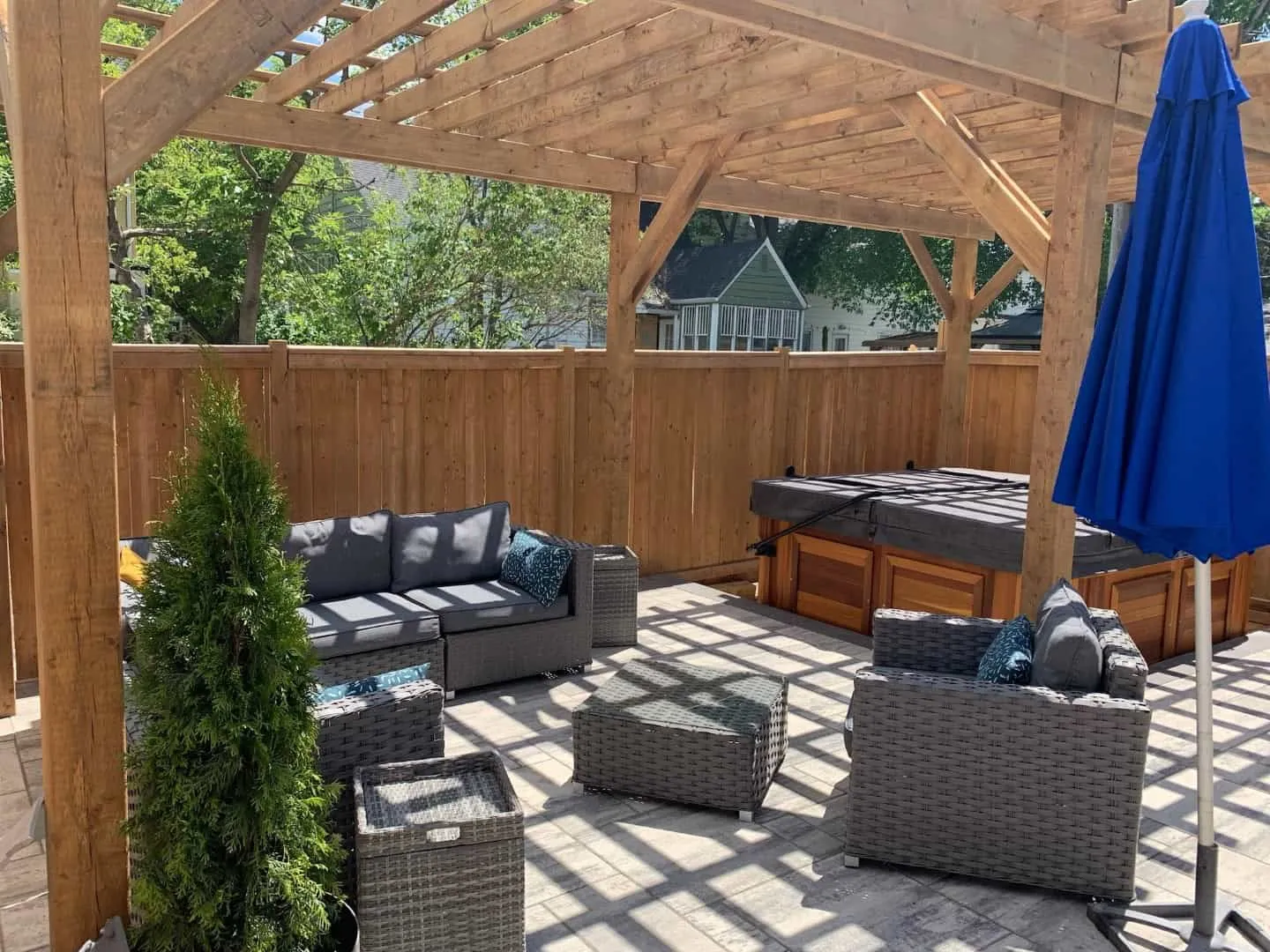 Grey paver patio under a timber pergola with wicker seating, a hot tub and a cedar fence