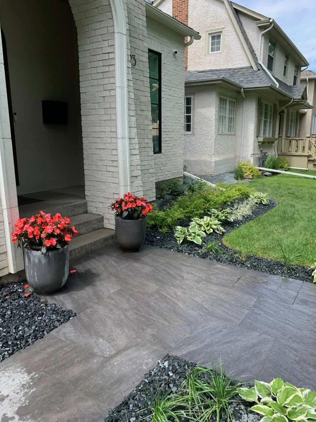 Large-format grey paver walkway leading to the arched brick entry of a house, edged with black rock and hosta beds