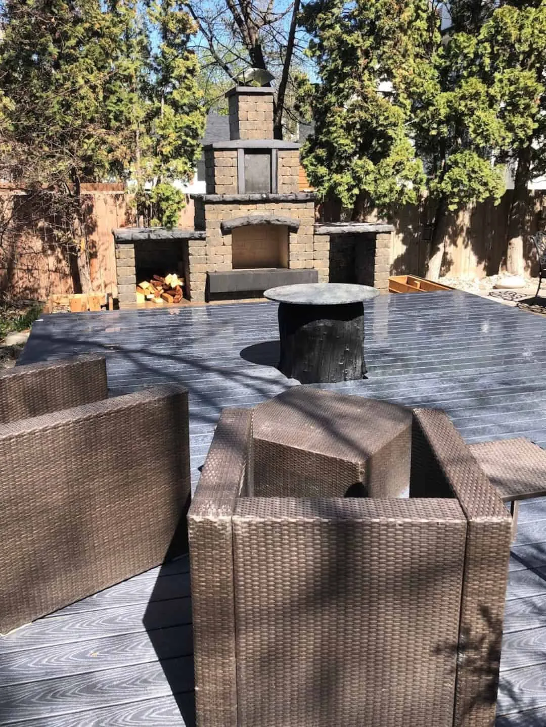 Block outdoor fireplace with a chimney and a firewood store built into its base, standing behind a composite deck with cedars either side