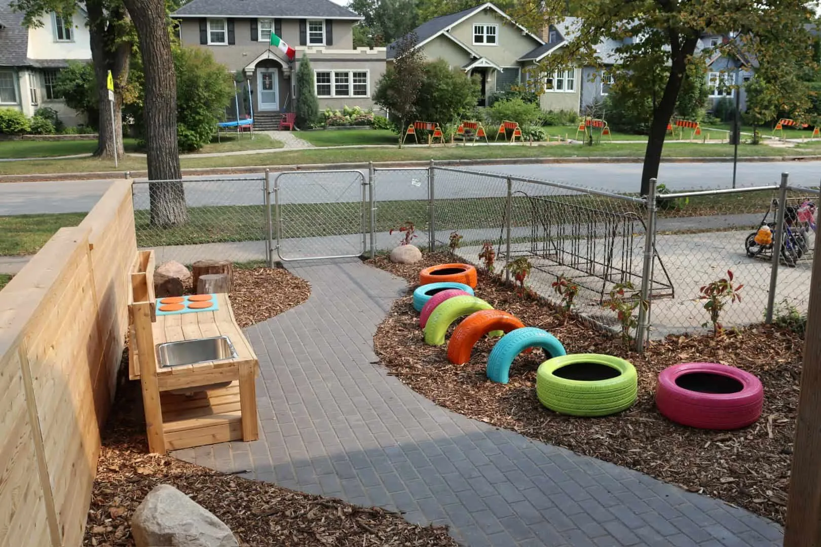 Charcoal paver walk between two beds of natural wood chip mulch at a daycare playground, with a cedar mud-kitchen bench and a boulder on one side and painted tyres on the other, and a chain-link gate to the street