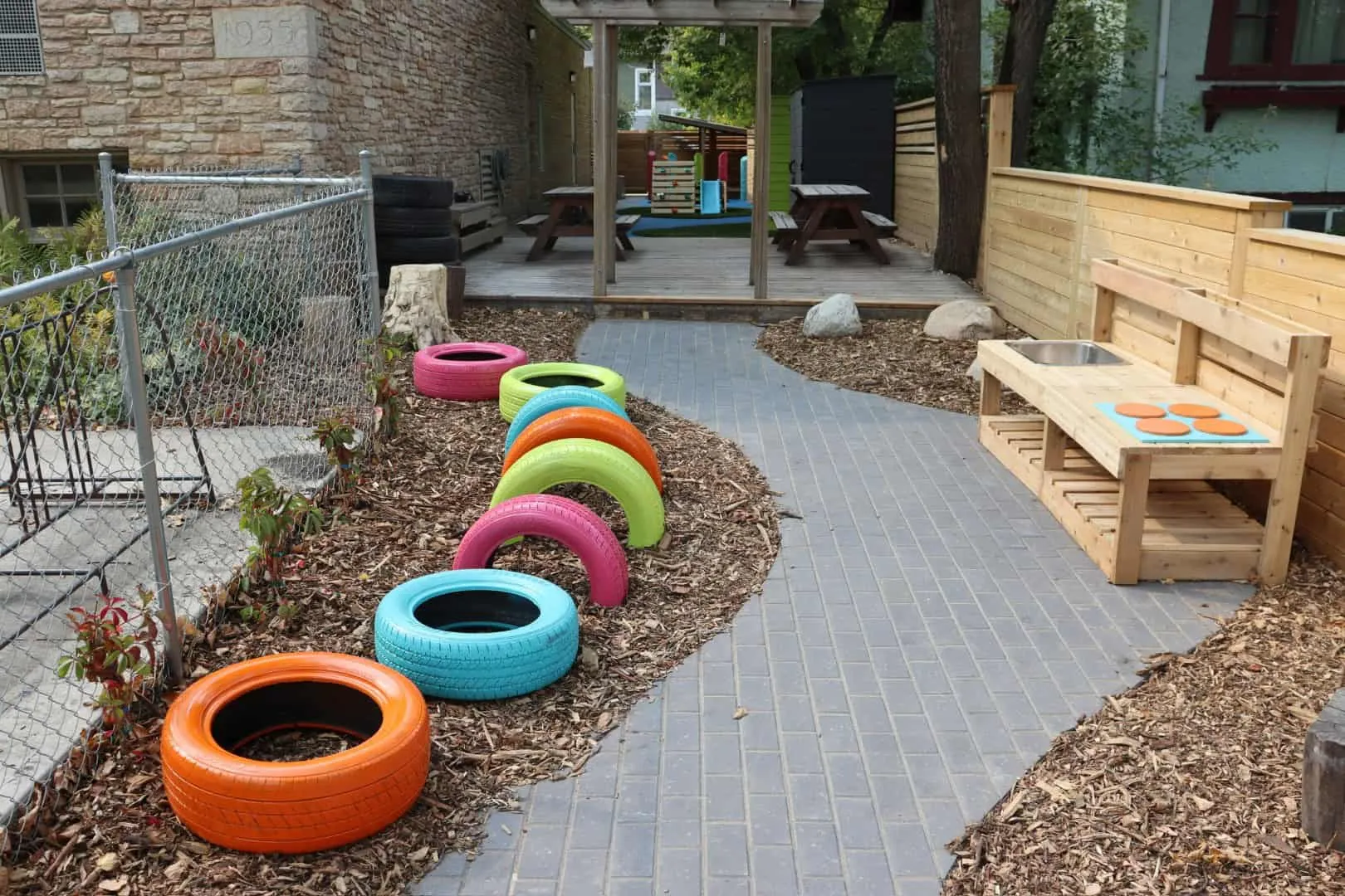 Charcoal paver walk between two beds of natural wood chip mulch at a daycare playground, with a cedar mud-kitchen bench and painted tyres set in the mulch